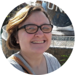 Mandy with shoulder length brown hair blowing in the wind, is wearing black eyeglasses and a gray shirt, is smiling at the camera.