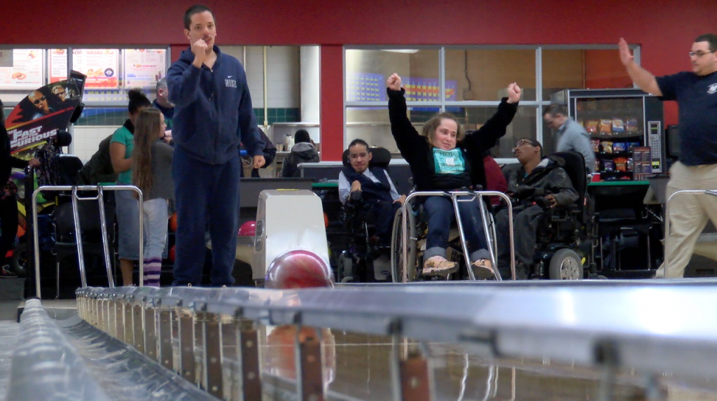 Group of people going bowling. Man on left is watching beyond the camera. Woman in wheelchair on right is raising her arms as if her bowling ball hit the pins.