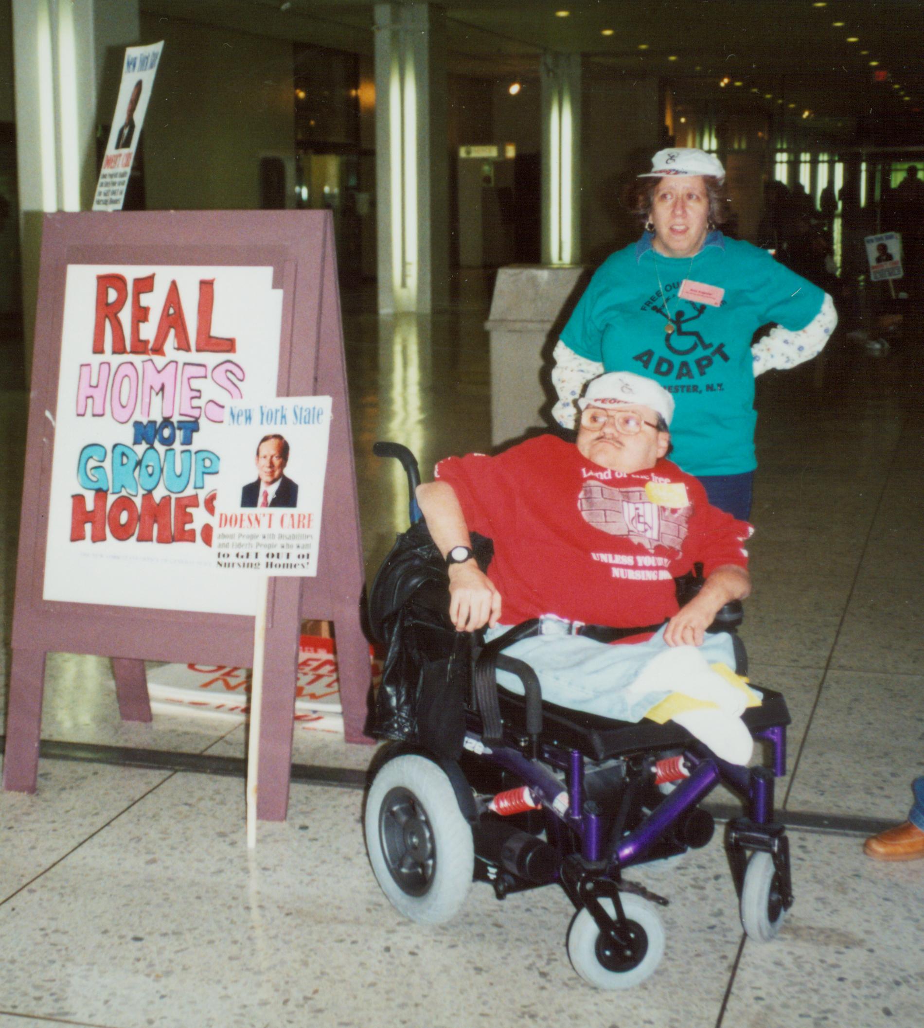 Picture of Joe Bonomo in his wheelchair next to a sign that reads 'Real Homes not Group Homes"